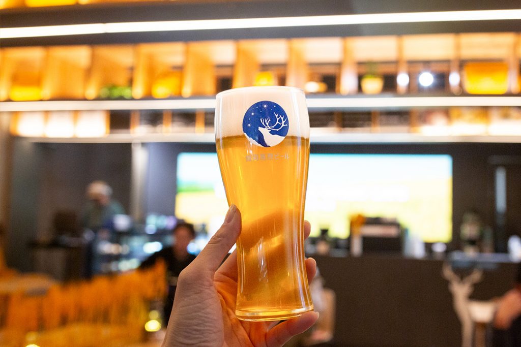 温かい光に包まれたカフェ店内で銀河高原ビールが注がれた銀河高原ビール専用グラスを手に持っている
